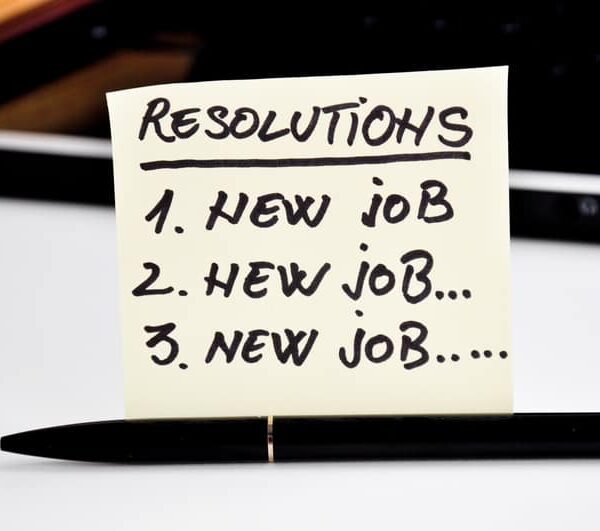 Yellow sticky note in front of a laptop reading Resolutions, 1 new job, 2 new job, 3 new job, with a black pen