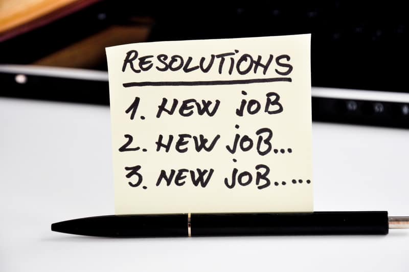 Yellow sticky note in front of a laptop reading Resolutions, 1 new job, 2 new job, 3 new job, with a black pen