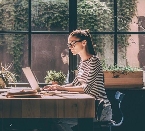Young woman in striped top working on a laptop at a wooden cafe table in front of a large window of climbing plants