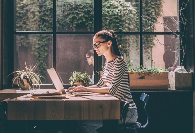 Young woman in striped top working on a laptop at a wooden cafe table in front of a large window of climbing plants