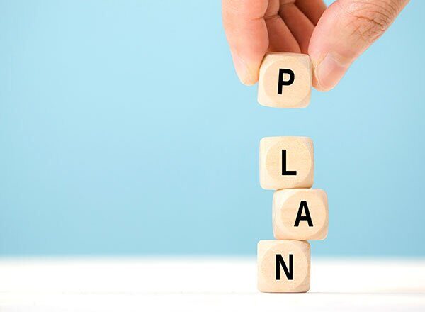 Hand placing a wooden block letter P on top of stacked blocks spelling LAN, forming the word PLAN against a blue background