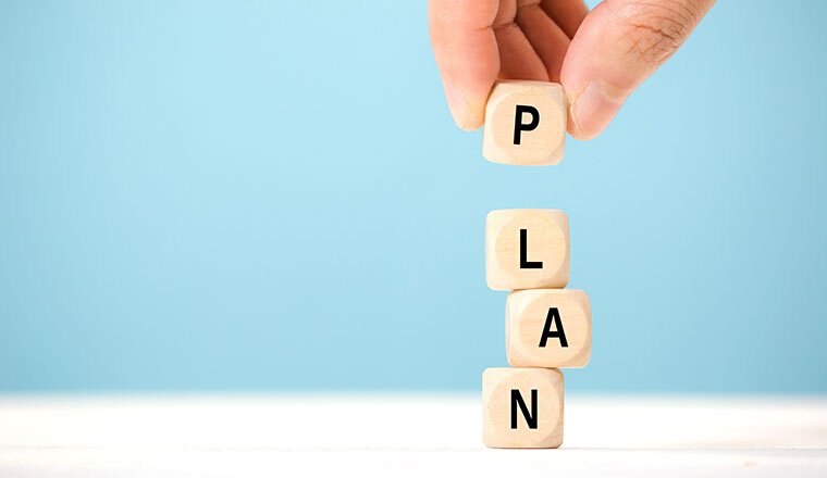 Hand placing a wooden block letter P on top of stacked blocks spelling LAN, forming the word PLAN against a blue background