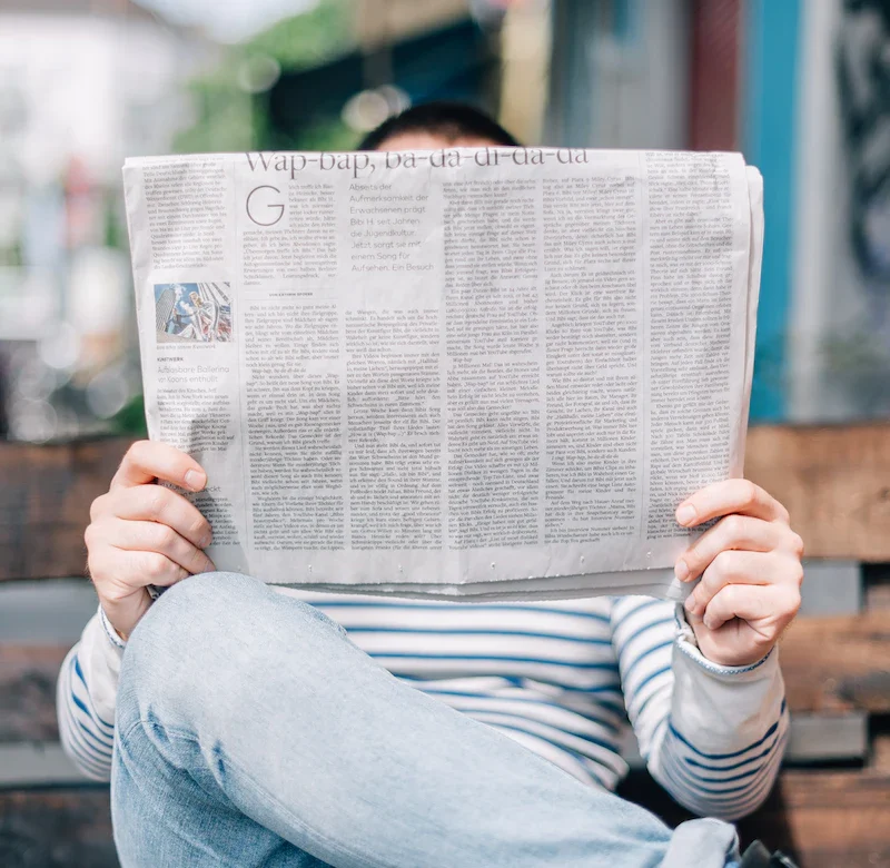 Person in striped shirt sitting on a wooden bench in jeans reading a large open newspaper, face obscured by the paper