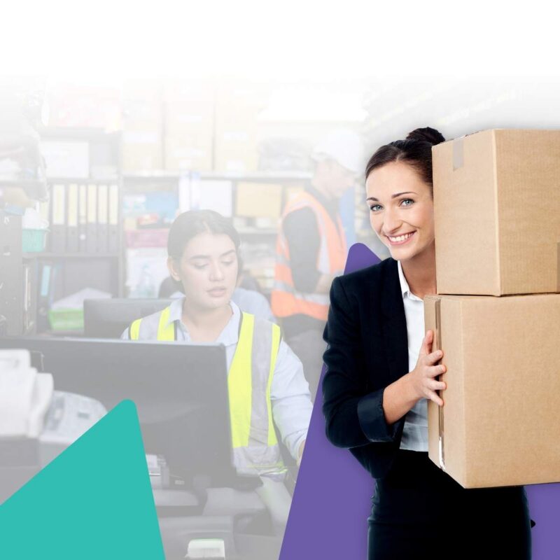 Smiling woman in blazer carrying stacked cardboard boxes beside a warehouse worker in hi-vis at a desk and a colleague in orange vest